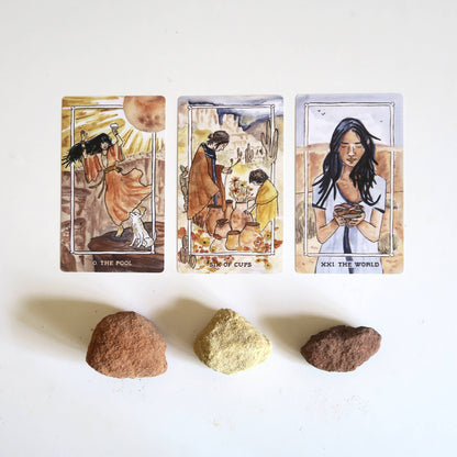 Three tarot cards with artistic illustrations of people and nature, accompanied by three stones on a white background.