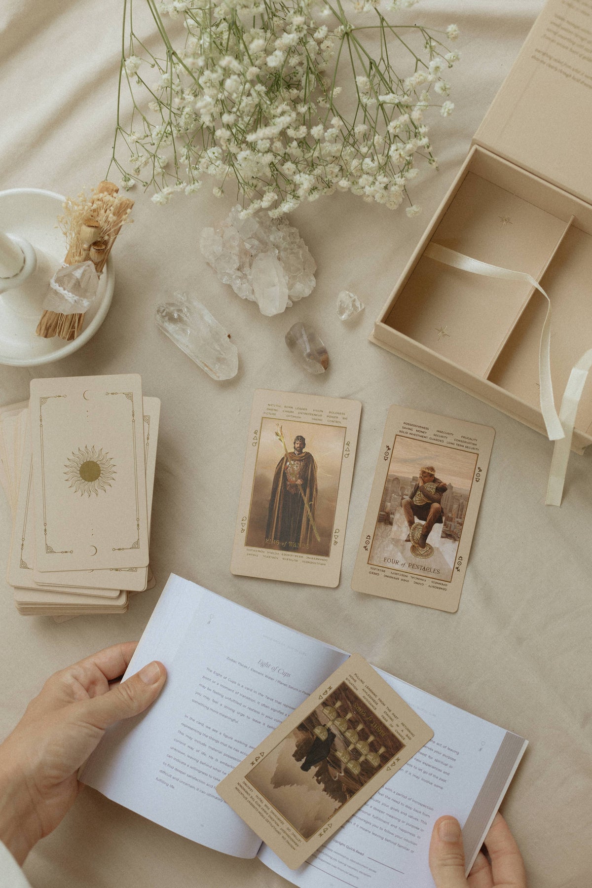 A styled flat-lay of a tarot card deck with an open booklet, several cards laid out, and hands holding an open page. White baby's breath flowers, crystals, a small dish, and an open gift box are arranged on a beige fabric background. Lucid Dreams Beginner's Tarot Beige Edition IV by ST. SOLEIL with gold foil cards, matte gold edges and guidebook