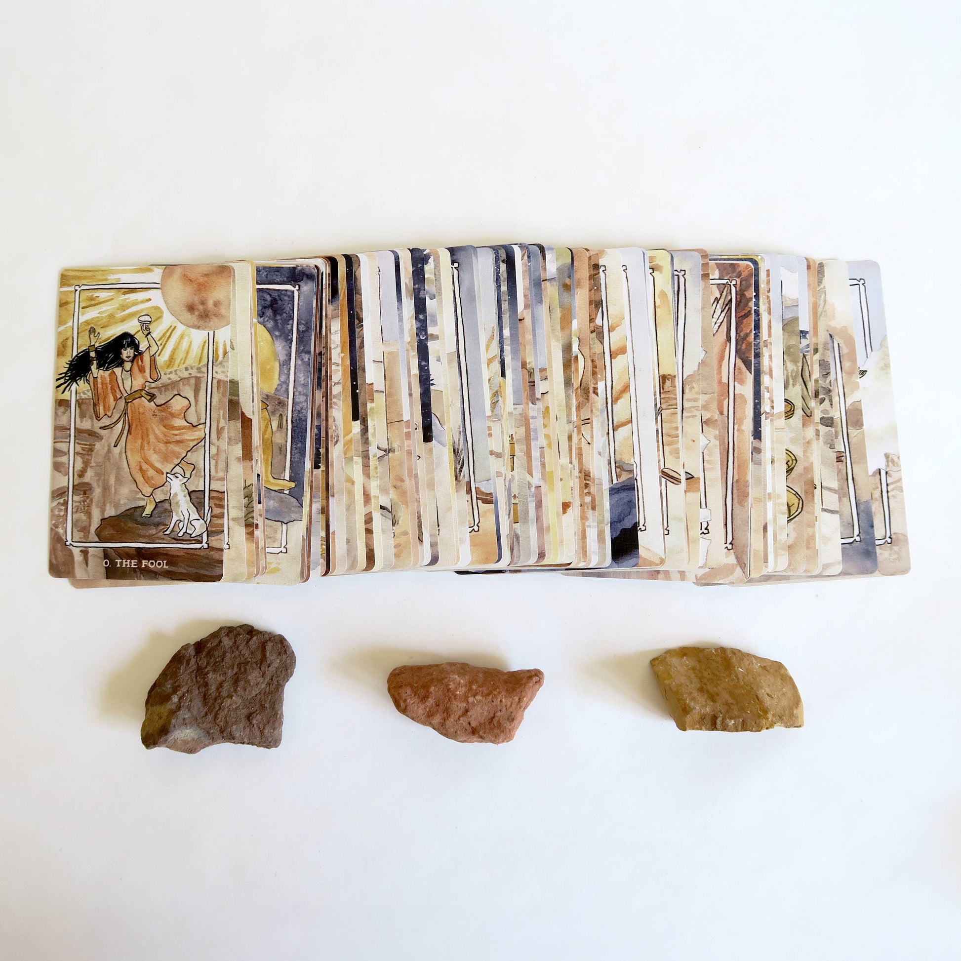 A fan of illustrated tarot cards spread out on a white surface, with three small brown stones placed below them.