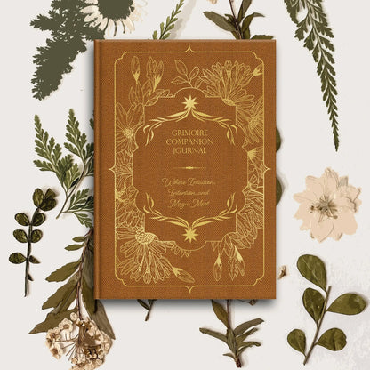 Brown 'Grimoire Companion Journal' surrounded by green leaves and flowers on a white background Grimoire Bouchette and Co Oneness Emporium