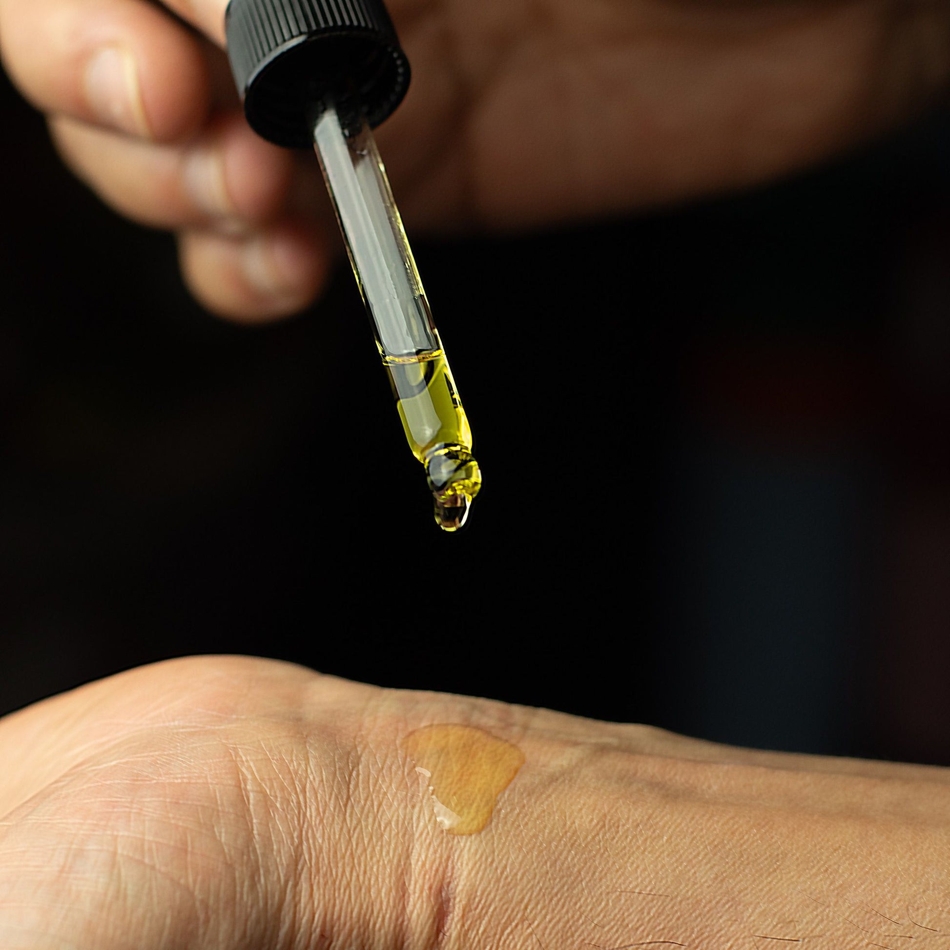 Hand holding a dropper with yellow liquid above a small patch on skin against a dark background