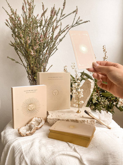Hand holding a card over books and decorative items on a white surface with flowers in the background Lucid Dreams Beginner's Tarot Beige Edition IV by ST. SOLEIL with gold foil cards, matte gold edges and guidebook