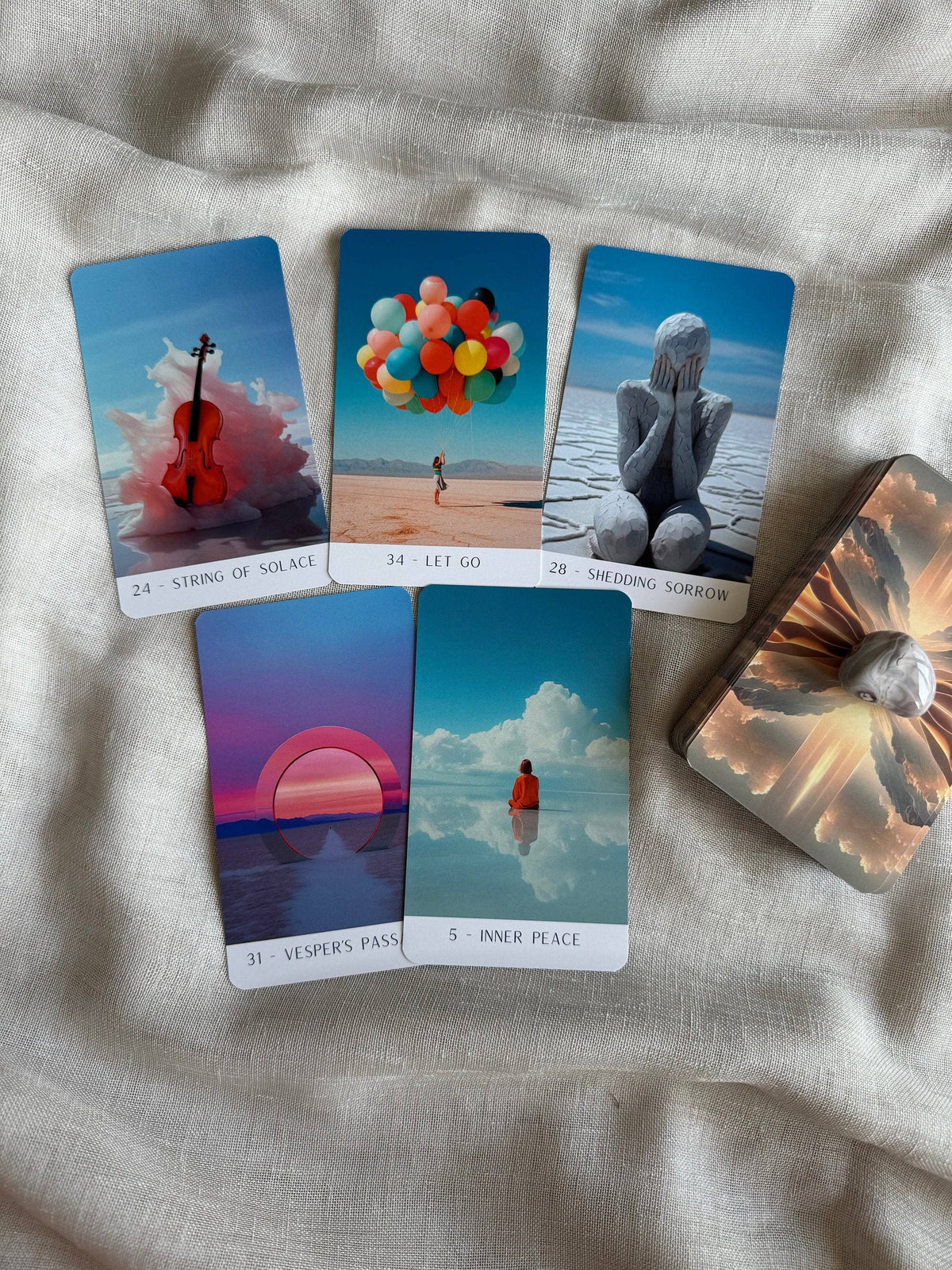 A set of illustrated oracle or tarot cards arranged on a light beige fabric surface, with the card deck box visible on the right. The cards show surreal scenic artwork: a violin in clouds, balloons on a beach, a stone figure by water, a sunset over water, and a reflective landscape with a small figure.