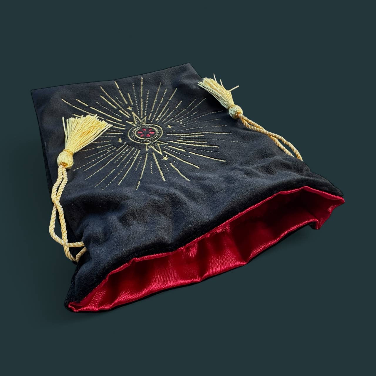 A velvet pouch with a dark navy exterior, gold sunburst embroidery, and a red satin lining, featuring cream-colored tassels and cords, shown on a dark teal background.