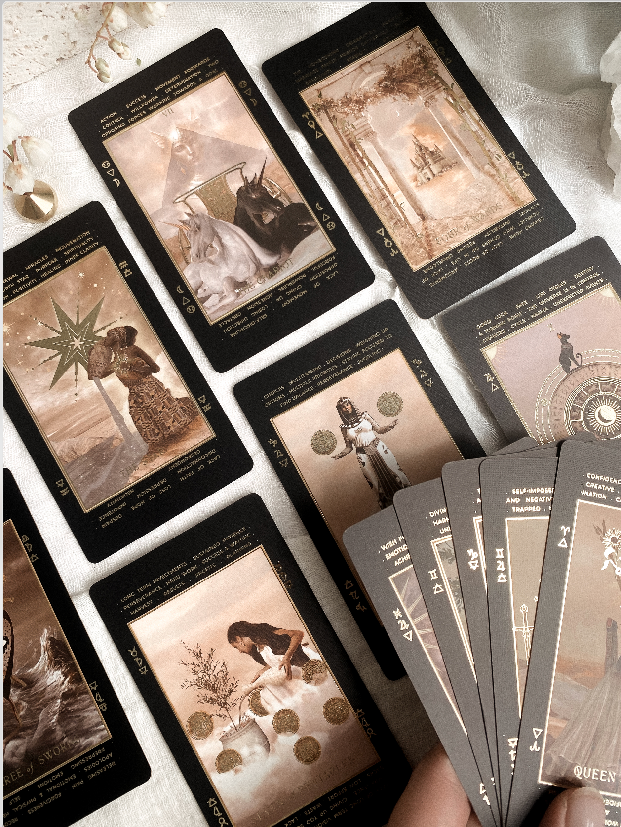 A set of tarot or oracle cards arranged on a light fabric surface, with a hand holding a fan of cards in the lower right corner. The cards have dark borders and sepia-toned illustrated scenes.