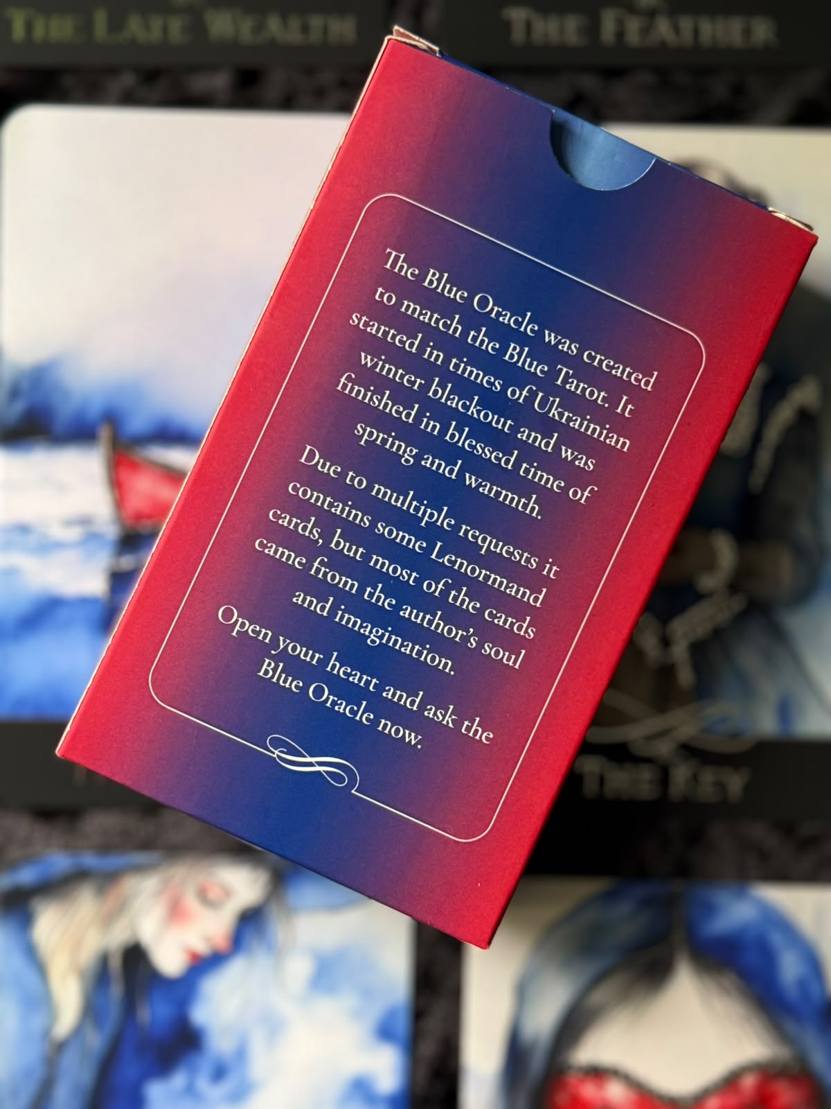The Blue Oracle card deck box shown from the back, featuring a red-to-blue gradient cover with white text, photographed against a blurred card-art background.