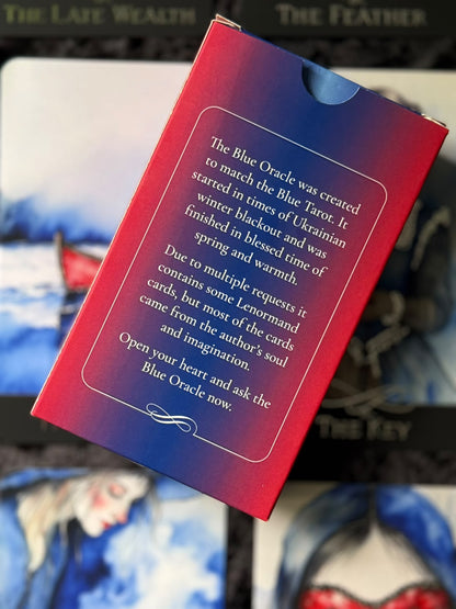 The Blue Oracle card deck box shown from the back, featuring a red-to-blue gradient cover with white text, photographed against a blurred card-art background.