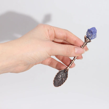 Hand holding a decorative spoon with a purple stone against a light background