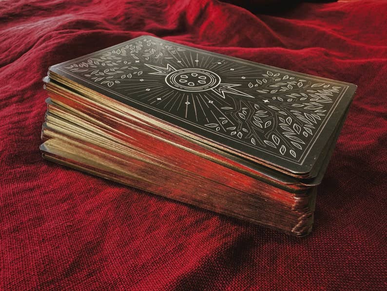 A deck of tarot cards with an ornate engraved black cover featuring a sun and leaf motifs, resting on a textured red fabric background. Asphodelon Tarot by Asphodelon Studio featuring hand-painted Greek mythology artwork, gilded edges, premium matte cards and a deluxe gold foil presentation box.