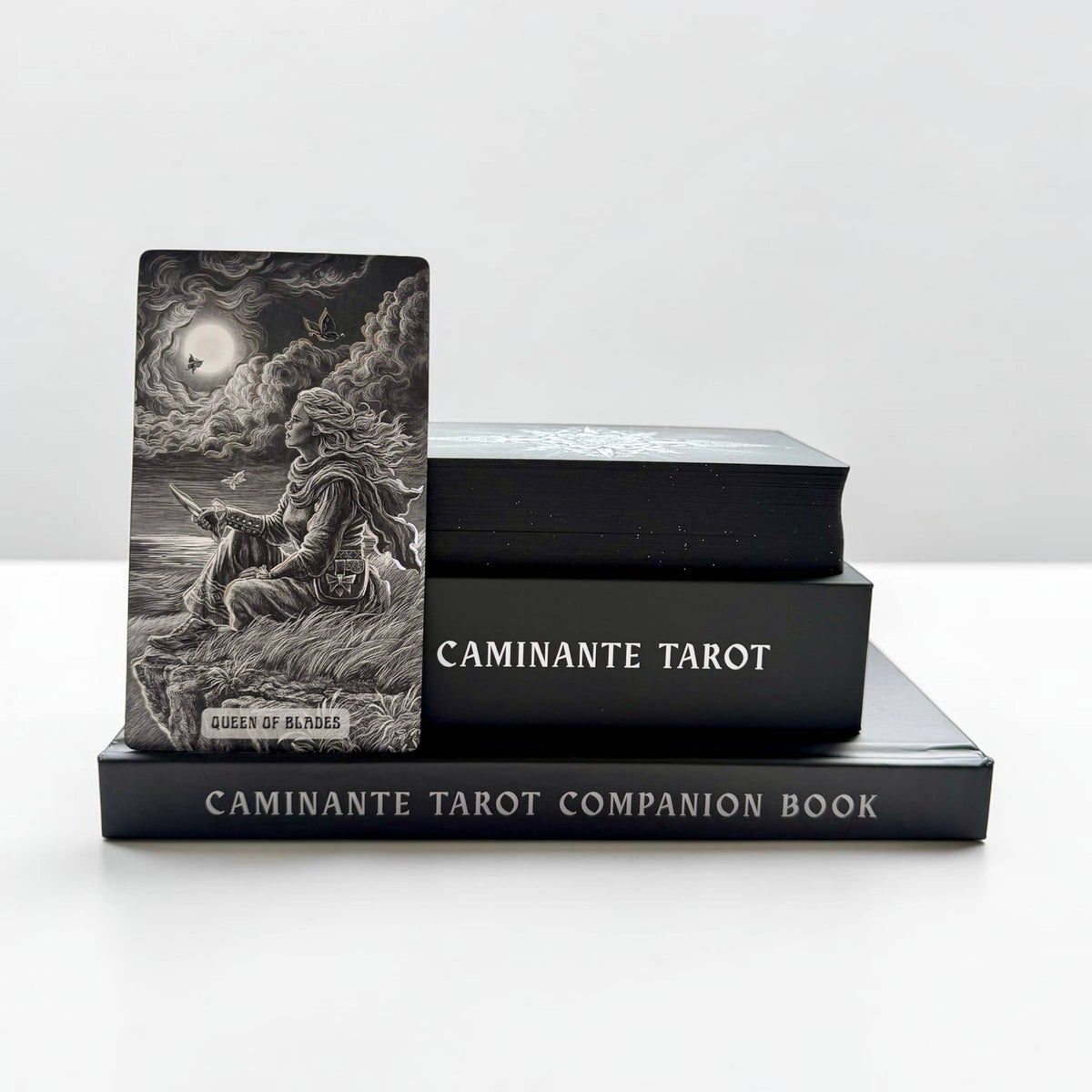 A tarot deck standing upright on a black companion book, with two more black tarot boxes stacked behind it on a white surface against a light gray background. The upright card shows a monochrome illustration of a seated figure in a stormy scene with a moon and clouds, and the visible text includes 'CAMINANTE TAROT' and 'CAMINANTE TAROT COMPAÑION BOOK'.
