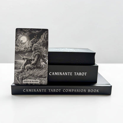 A tarot deck standing upright on a black companion book, with two more black tarot boxes stacked behind it on a white surface against a light gray background. The upright card shows a monochrome illustration of a seated figure in a stormy scene with a moon and clouds, and the visible text includes 'CAMINANTE TAROT' and 'CAMINANTE TAROT COMPAÑION BOOK'.