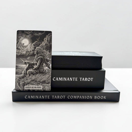 A tarot deck standing upright on a black companion book, with two more black tarot boxes stacked behind it on a white surface against a light gray background. The upright card shows a monochrome illustration of a seated figure in a stormy scene with a moon and clouds, and the visible text includes 'CAMINANTE TAROT' and 'CAMINANTE TAROT COMPAÑION BOOK'.