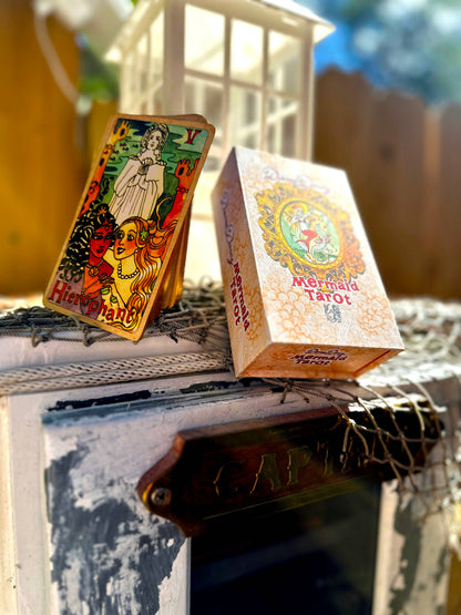 A boxed deck of Mermaid Tarot cards and one upright card displayed outdoors on a rustic white wooden surface with netting and a lantern in the background.