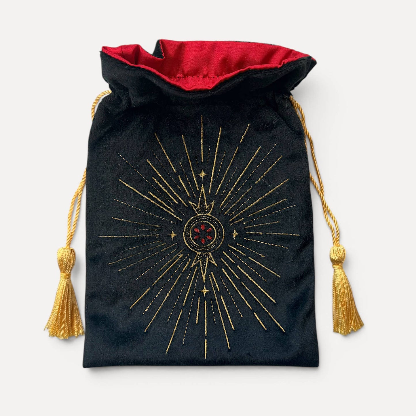 A black velvet drawstring pouch with a red velvet lining, featuring a gold embroidered sunburst design with a central red and gold emblem. The drawstrings are gold with matching tassels.