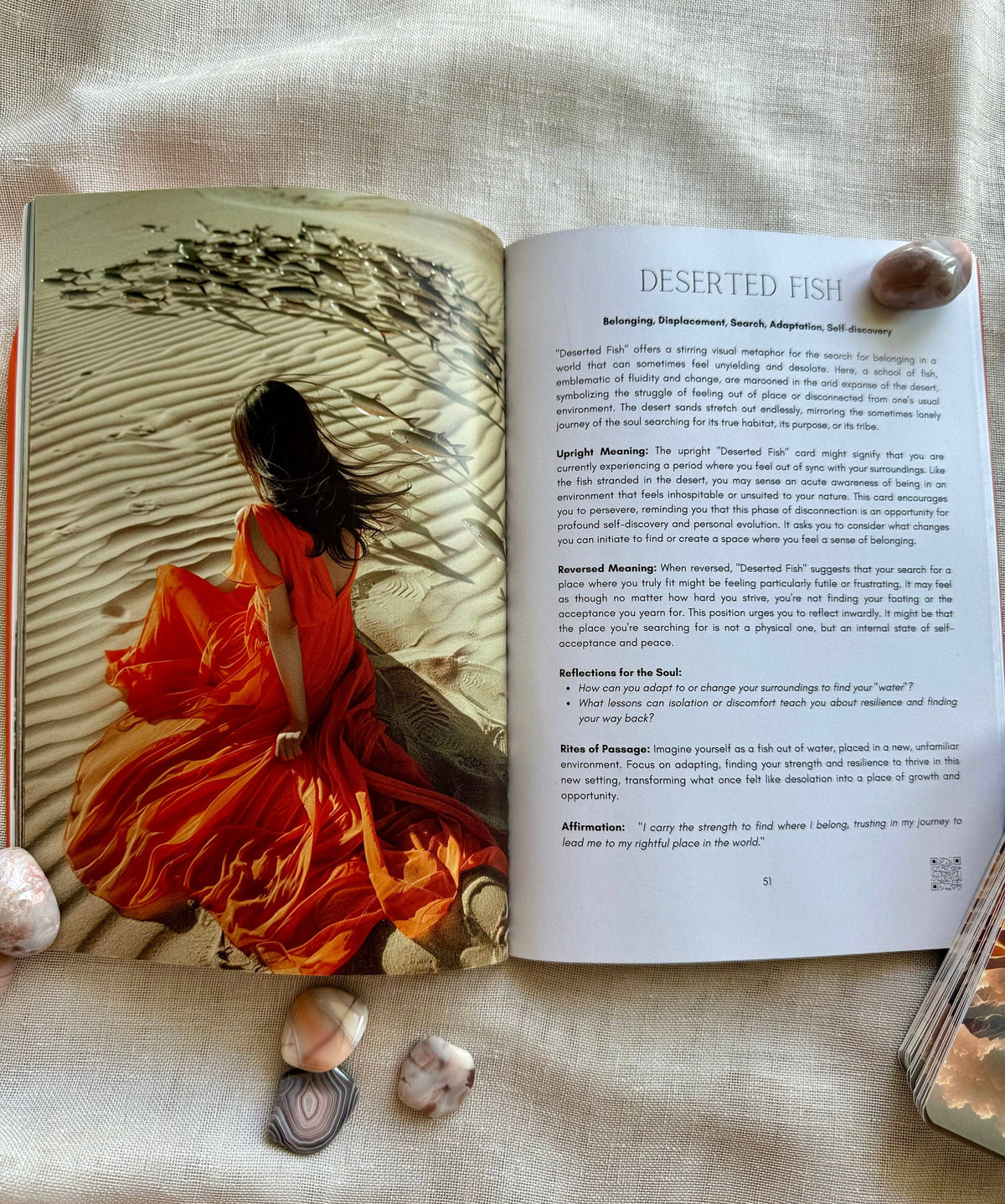 An open book spread laid on a light fabric background, showing a page titled 'DESERTED FISH' with text and a large photograph of a woman in a flowing orange robe seated among dunes. Several small shells are placed around the book.