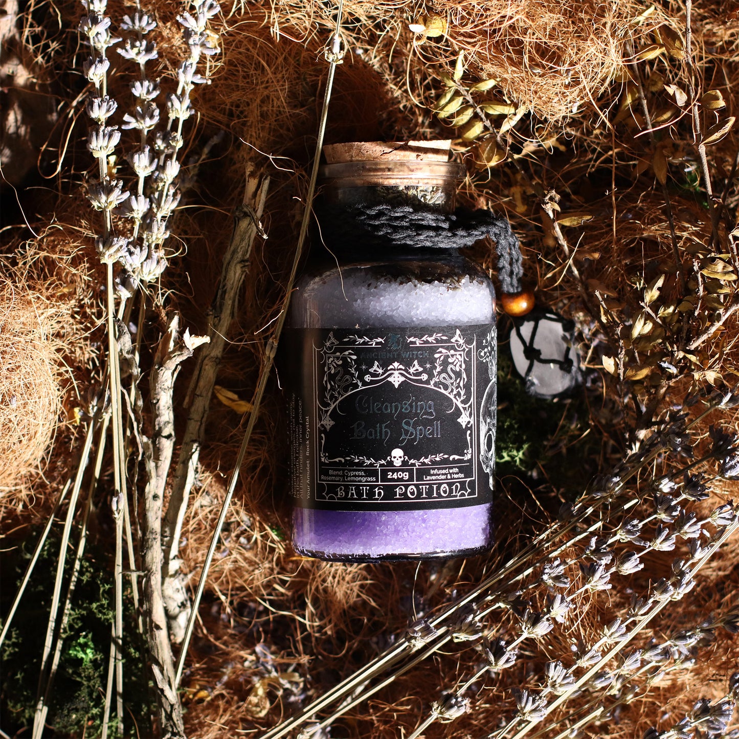 Purple bottle labeled 'Witching Bath Spell' surrounded by dried plants on a textured surface