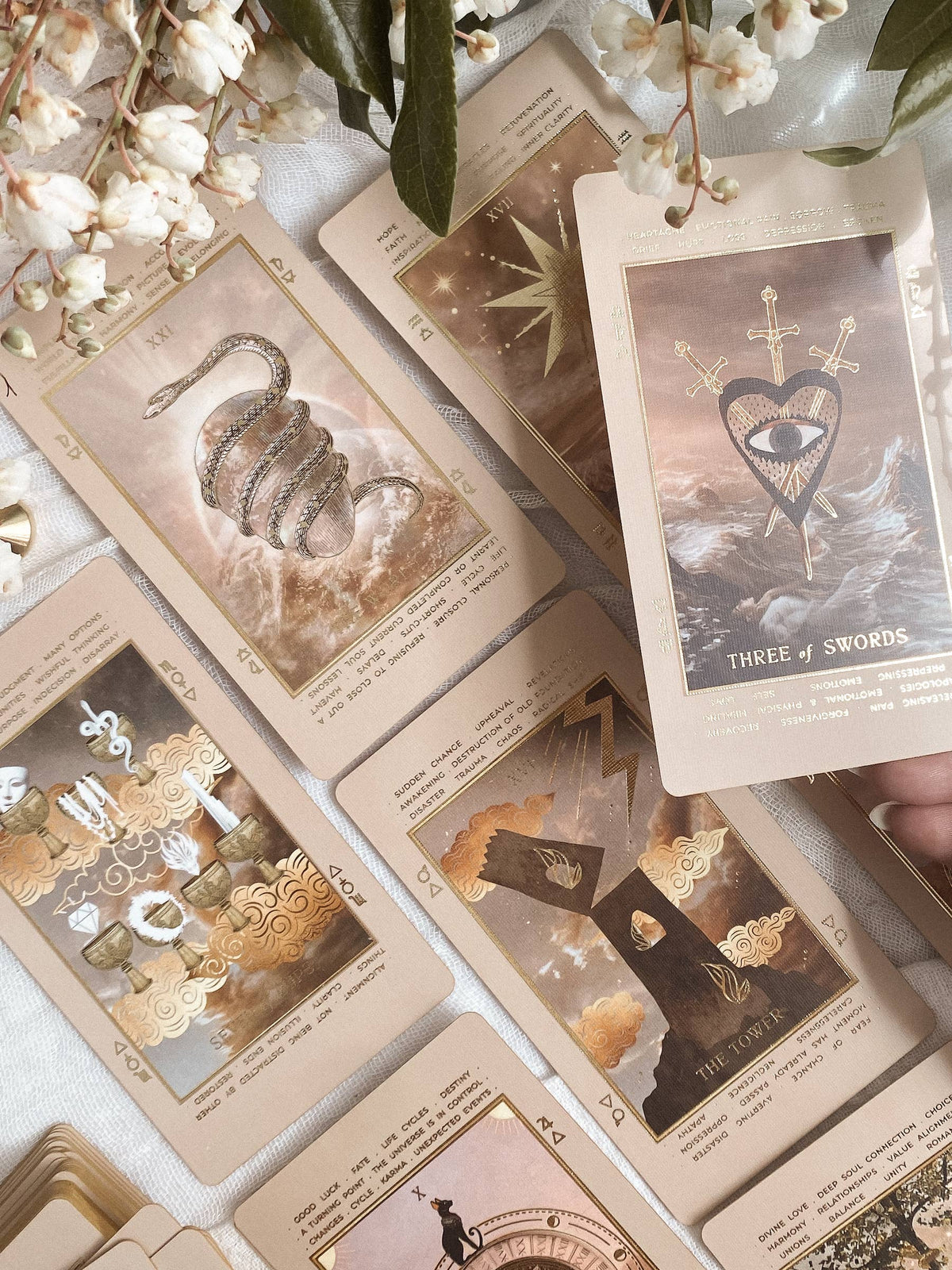 A styled flat-lay of beige oracle or tarot-style cards with illustrated and typographic artwork, arranged on a light fabric with white flowers and green leaves. A hand is holding one card at the right edge. Lucid Dreams Beginner's Tarot Beige Edition IV by ST. SOLEIL with gold foil cards, matte gold edges and guidebook