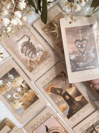 A styled flat-lay of beige oracle or tarot-style cards with illustrated and typographic artwork, arranged on a light fabric with white flowers and green leaves. A hand is holding one card at the right edge. Lucid Dreams Beginner's Tarot Beige Edition IV by ST. SOLEIL with gold foil cards, matte gold edges and guidebook