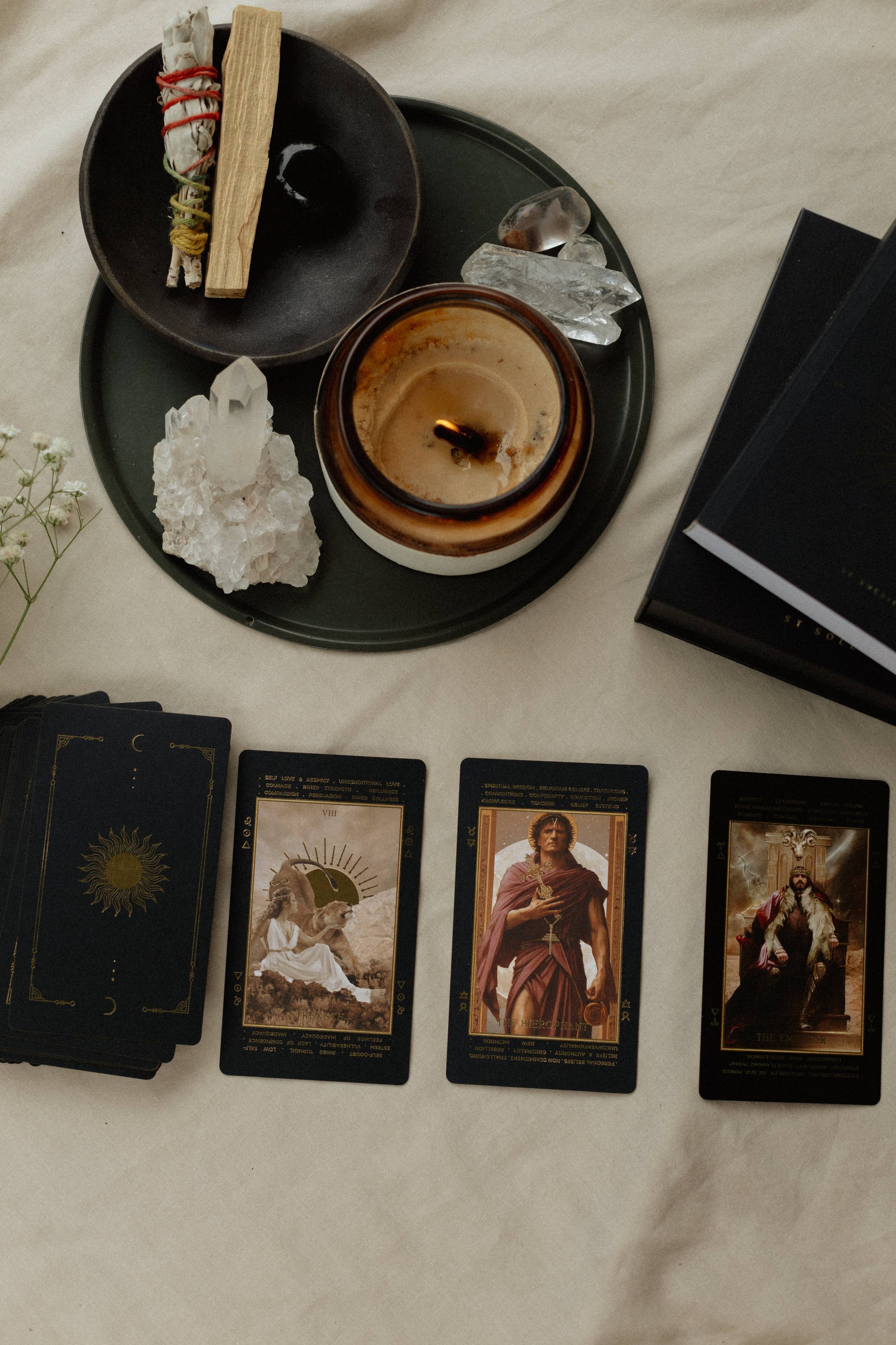 A styled flat-lay of tarot cards and ritual accessories on a cream fabric background. Four tarot cards are laid out in a row, alongside a deck of cards, a black candle, a smudge bundle, crystals, and a black book.