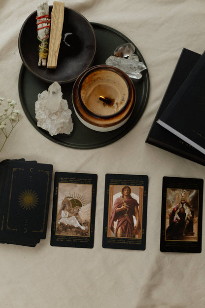 A styled flat-lay of tarot cards and ritual accessories on a cream fabric background. Four tarot cards are laid out in a row, alongside a deck of cards, a black candle, a smudge bundle, crystals, and a black book.