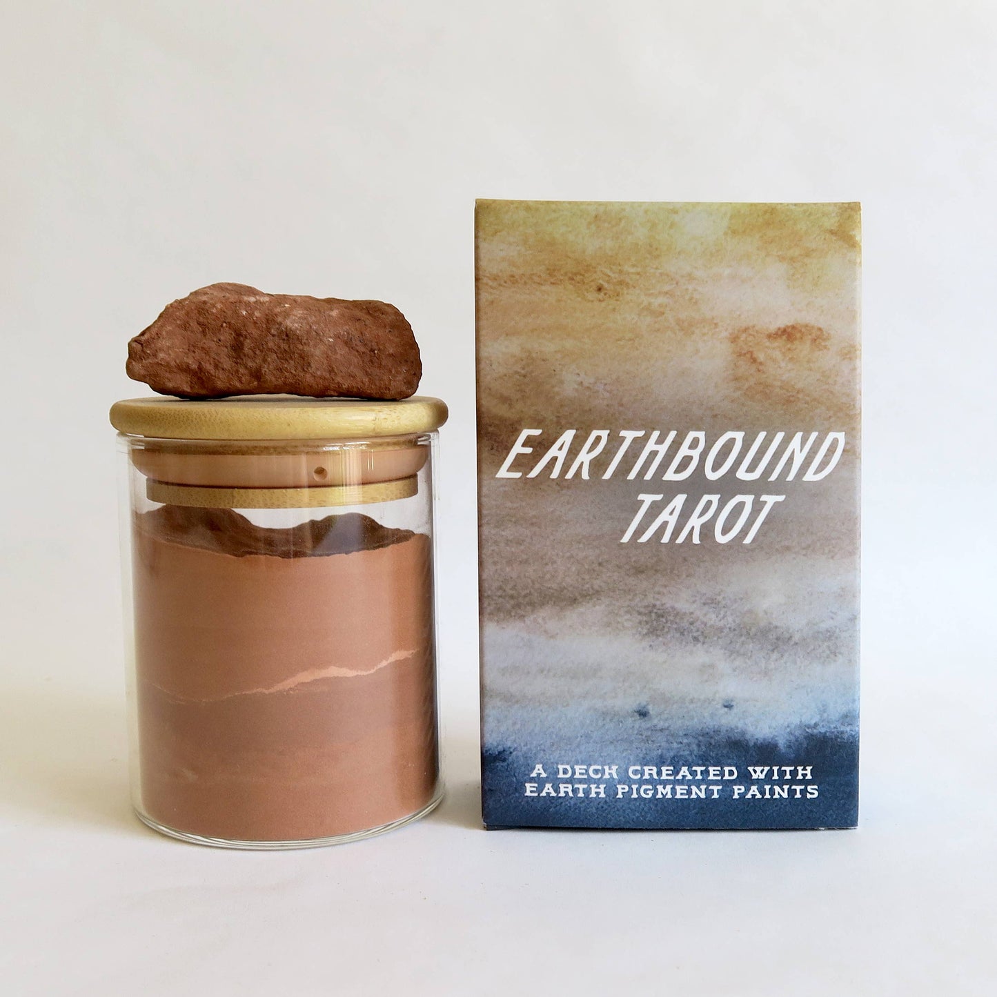 Earthbound Tarot deck shown with its product box and a glass jar containing brown earth pigment powder, topped with a small brown rock, on a clean white background.