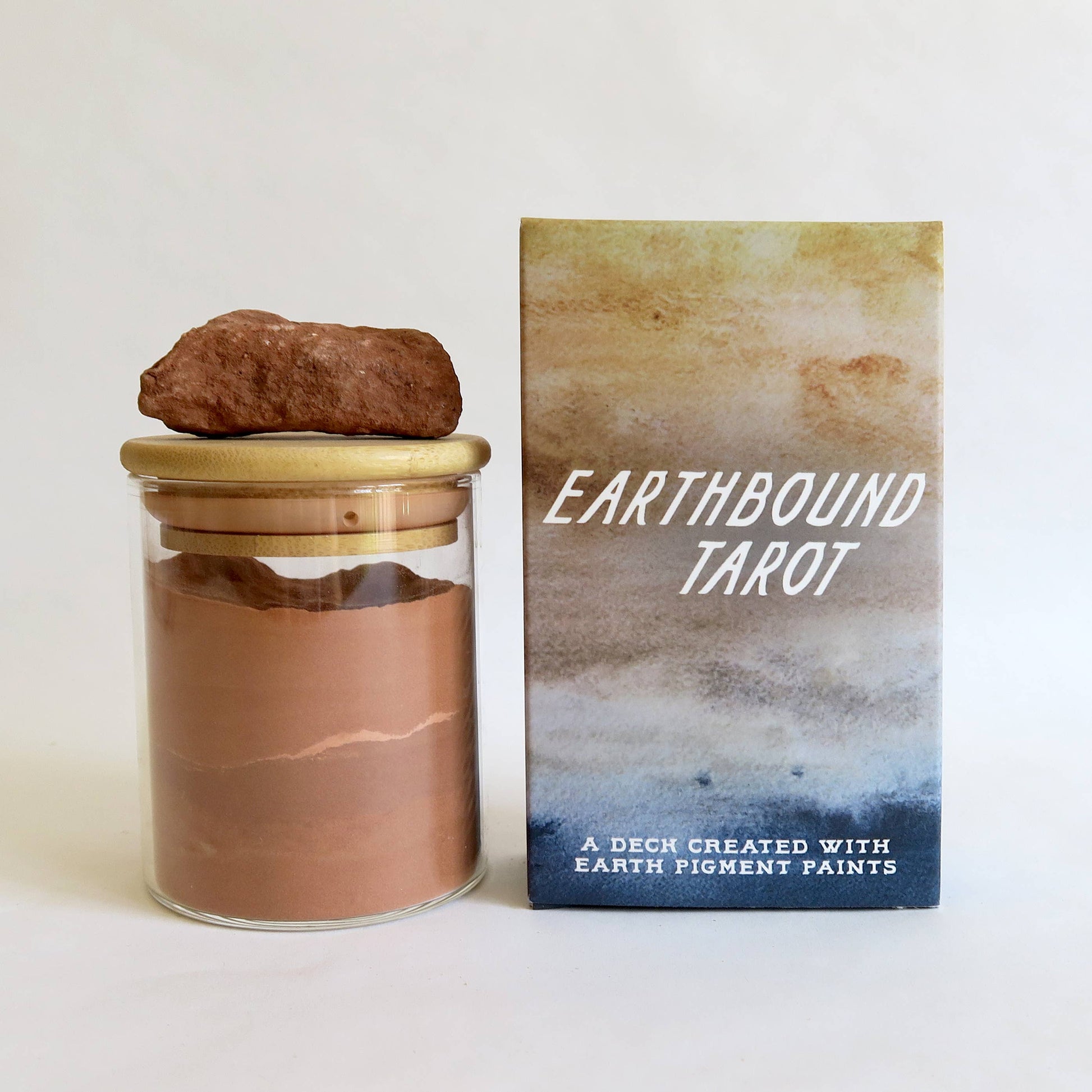 Earthbound Tarot deck shown with its product box and a glass jar containing brown earth pigment powder, topped with a small brown rock, on a clean white background.