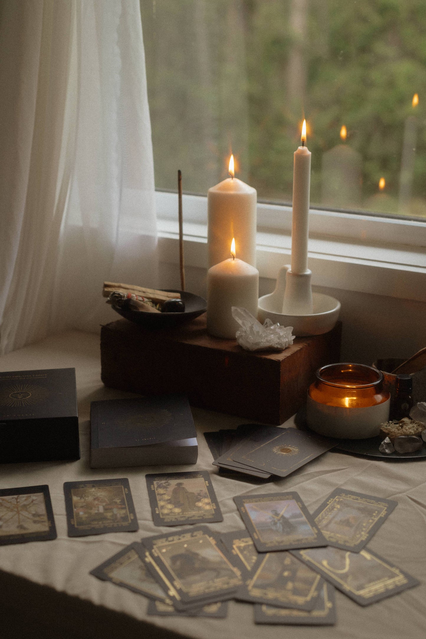 A styled tabletop scene by a window with lit white candles, tarot or oracle cards spread out, crystals, a small amber candle, and boxed items on a draped table. No person or animal is present, but this is not a clean isolated studio product shot; it is a real interior environment, so subject_type is Lifestyle.