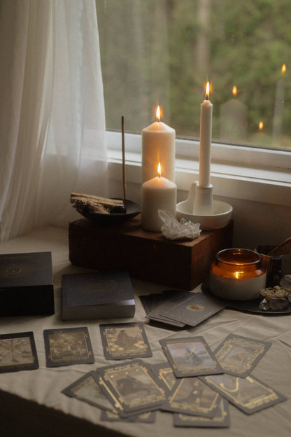 A styled tabletop scene by a window with lit white candles, tarot or oracle cards spread out, crystals, a small amber candle, and boxed items on a draped table. No person or animal is present, but this is not a clean isolated studio product shot; it is a real interior environment, so subject_type is Lifestyle.