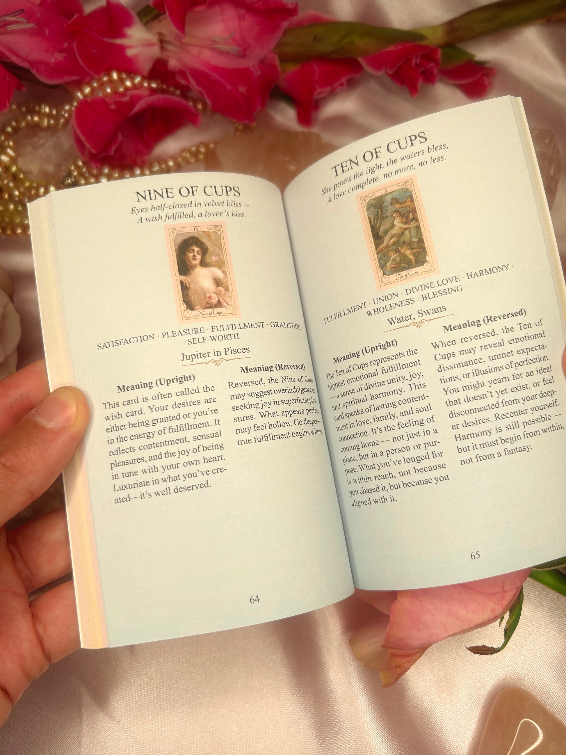 Open book with tarot card meanings, held against a soft pink background with flowers. Ethereal Goddess Tarot guidebook