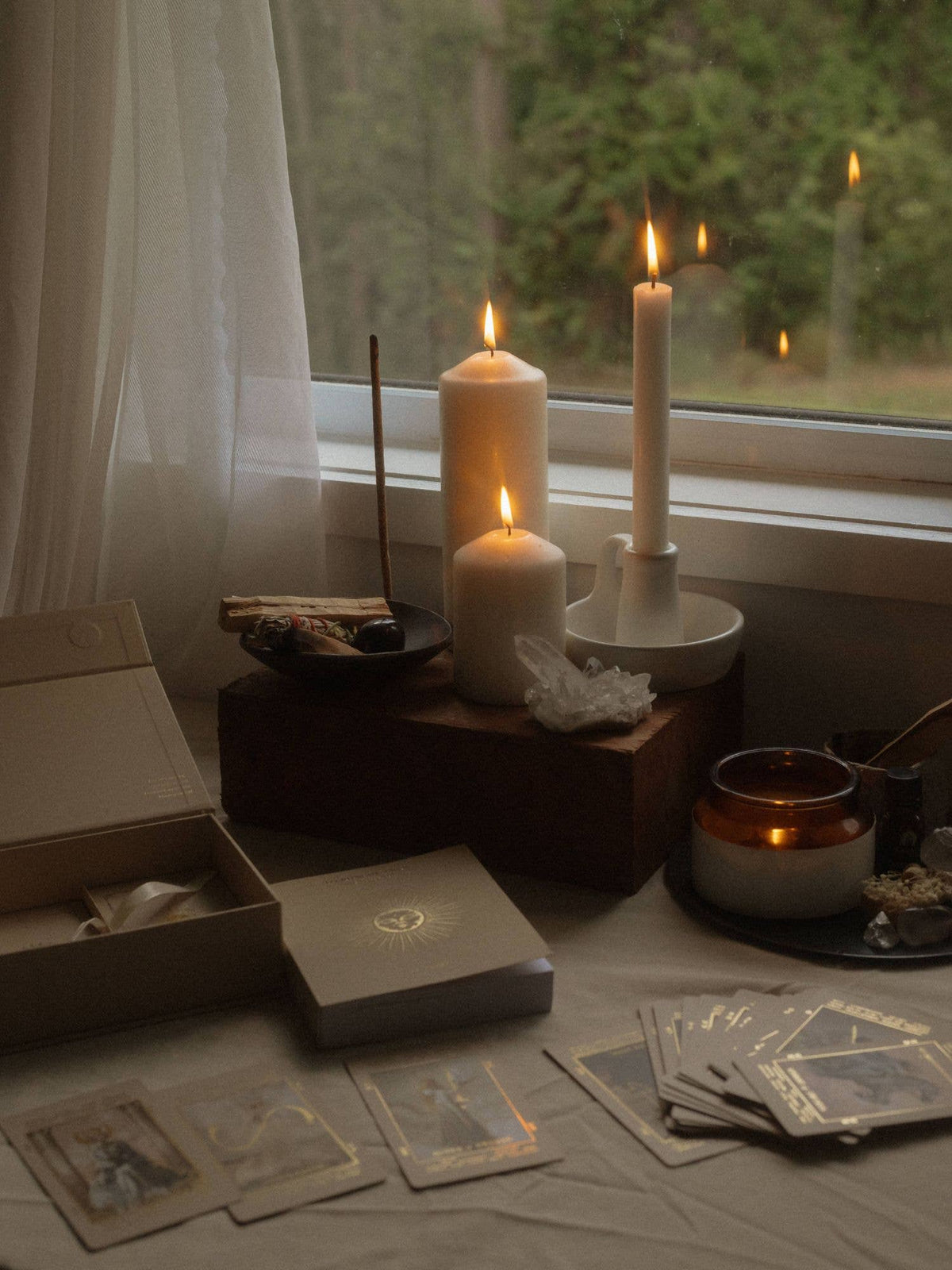 A styled arrangement of candles, tarot cards, crystals, and an open box on a table by a window. The scene is softly lit with warm candlelight, showing white pillar candles, a lit tealight candle, a small amber candle, and several illustrated cards. Lucid Dreams Beginner's Tarot Beige Edition IV by ST. SOLEIL with gold foil cards, matte gold edges and guidebook
