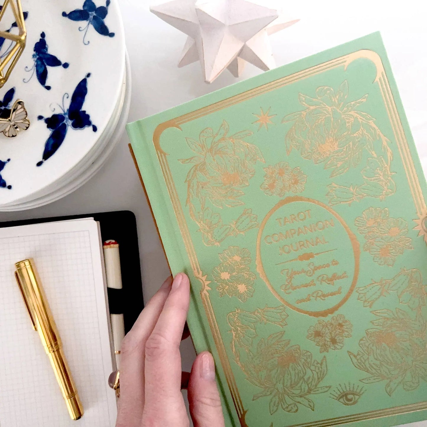 Decorative book with gold embossed design held by a hand, with a plate featuring blue butterflies in the background. Tarot Journal Bouchette and Co Oneness Emporium