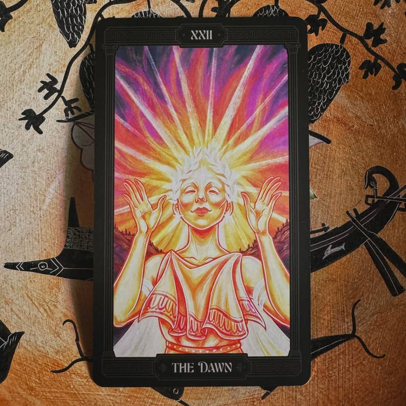 A tarot card titled 'The Dawn' (XXII) featuring a colorful illustration of a radiant figure with raised hands, set against a black border and placed on a patterned brown background. Asphodelon Tarot by Asphodelon Studio featuring hand-painted Greek mythology artwork, gilded edges, premium matte cards and a deluxe gold foil presentation box.