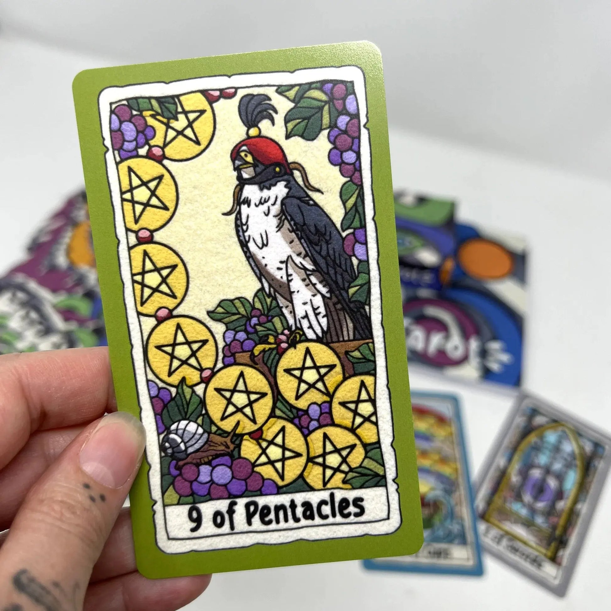 Card with a rooster and pentacle symbols held by a hand, with other cards in the background.