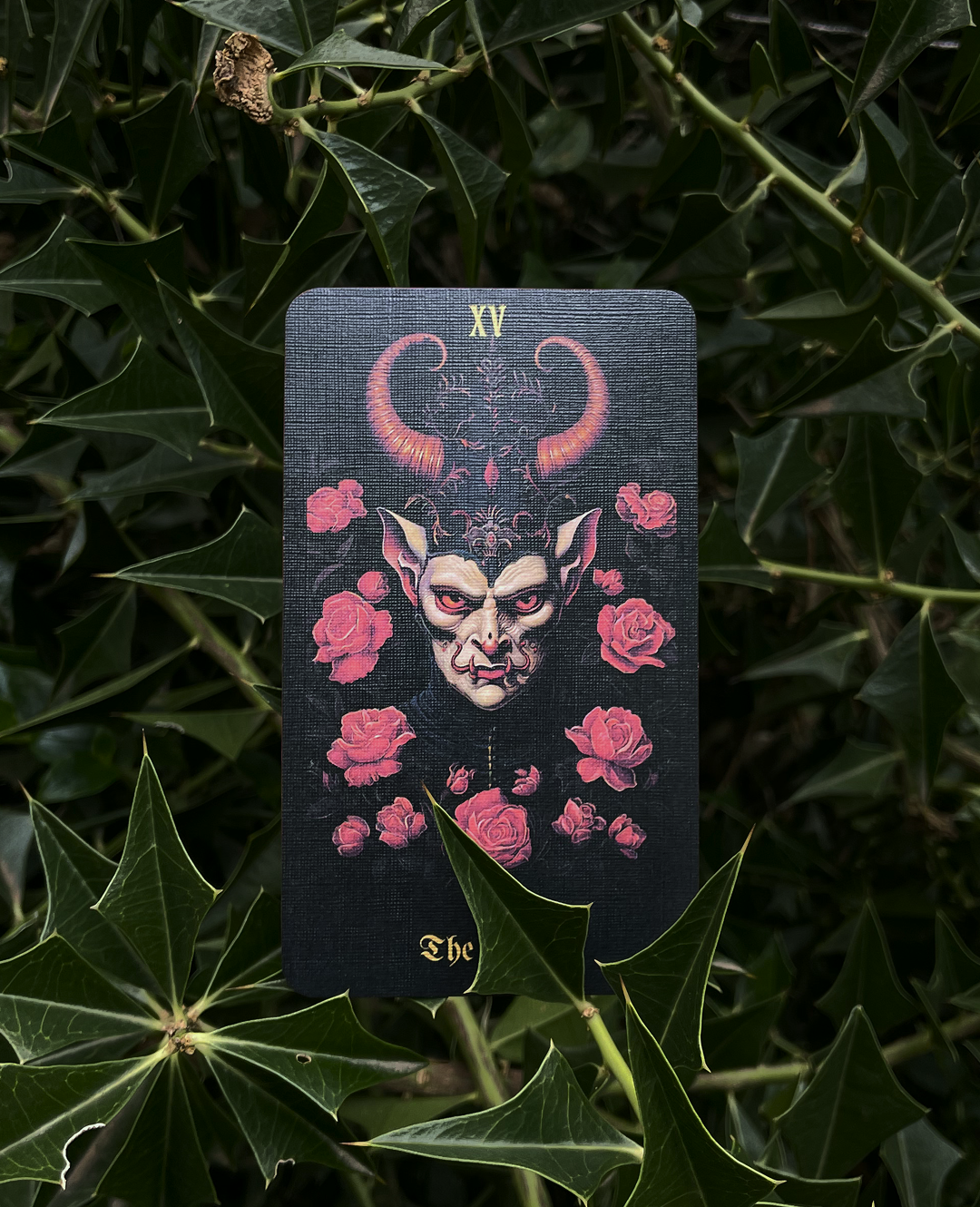 Tarot card with devil illustration and roses on a dark background, surrounded by green leaves.