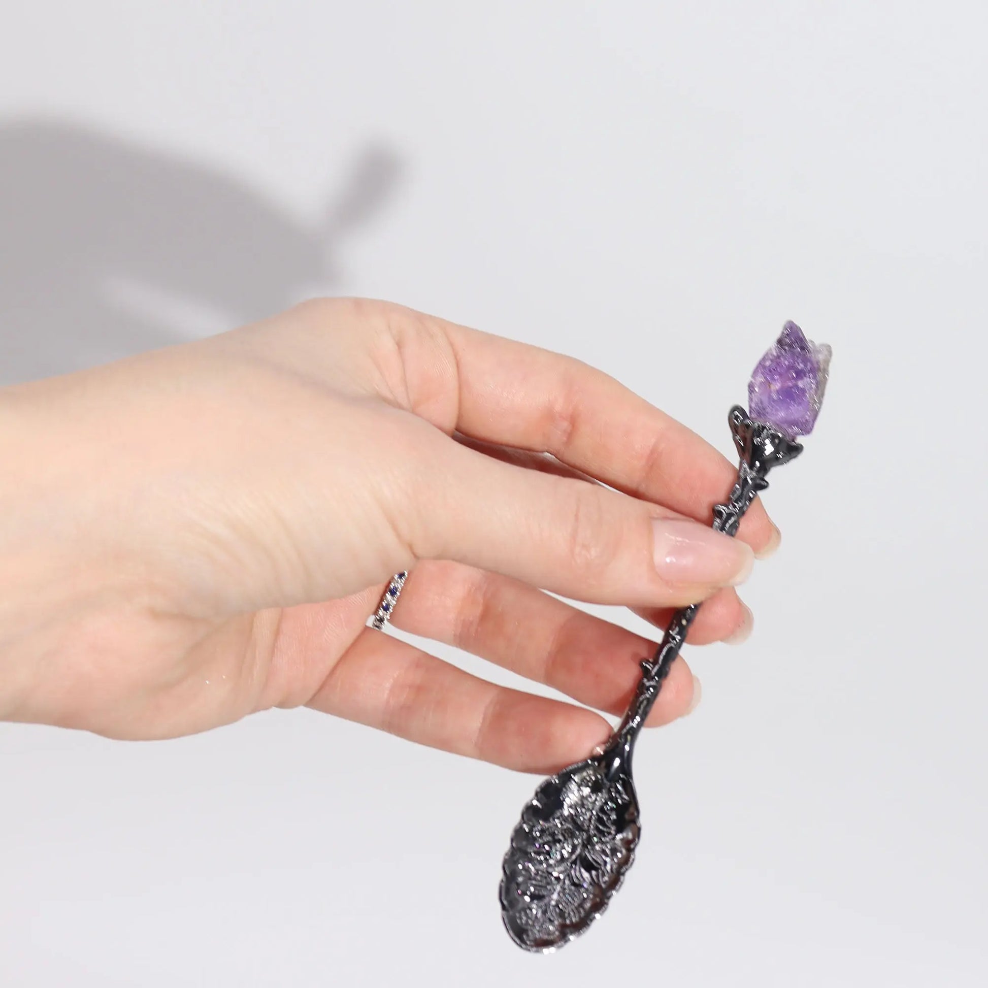 Hand holding a decorative spoon with a purple crystal on a light gray background