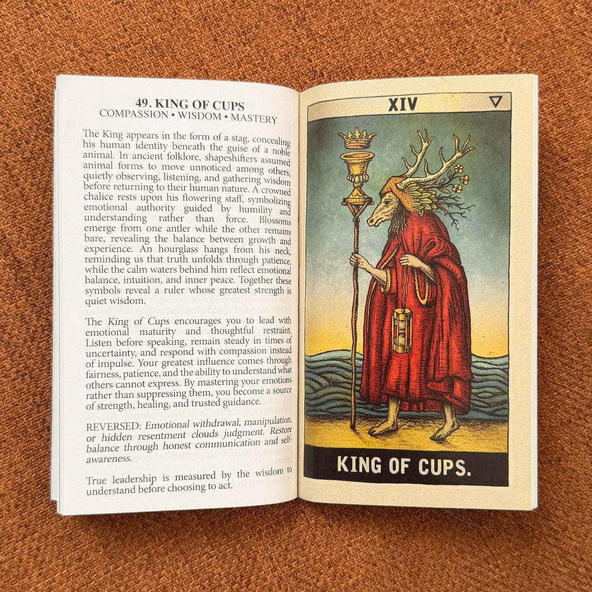A printed card or booklet page laid on a brown textured surface, showing a page with the heading '49. KING OF CUPS' and text on the left, plus an illustrated cup illustration on the right with the title 'KING OF CUPS.